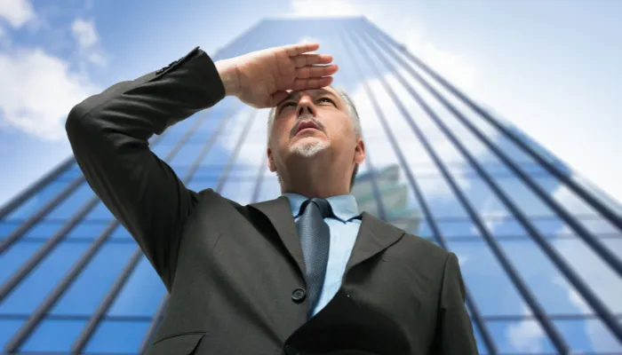 homem colocando a mão na cabeça, como se olhasse para o futuro, na frente de um prédio empresarial de uma grande empresa de seguros.