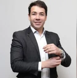 Johnatan Queiroz é o fundador da Lucma Seguros, hoje Grupo Lucma Seguros, nesta imagem ele está segurando a manga do terno com a mão direita, está usando relógio, terno de cor cinza e sorrindo para foto.