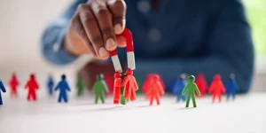 homem segurando ímã, puxando leads desqualificados, com bonecos representando leads, nas cores azul, verde e vermelho. O homem está sentado à uma mesa, com blusa de manga cumprida azul, o homem possui pele escura.