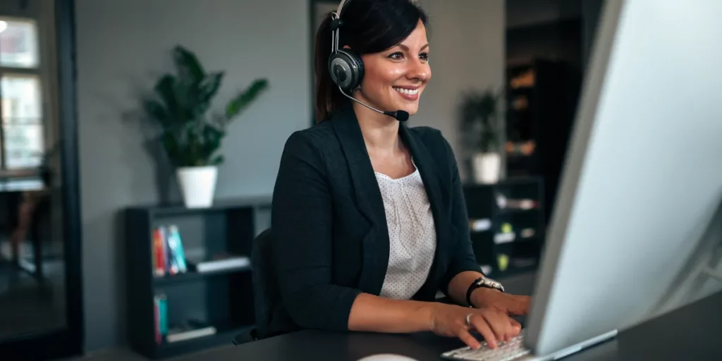 mulher com fone de ouvido sorrindo, em frente ao computador, falando com seus clientes, para venda de carteira de clientes de seguros.